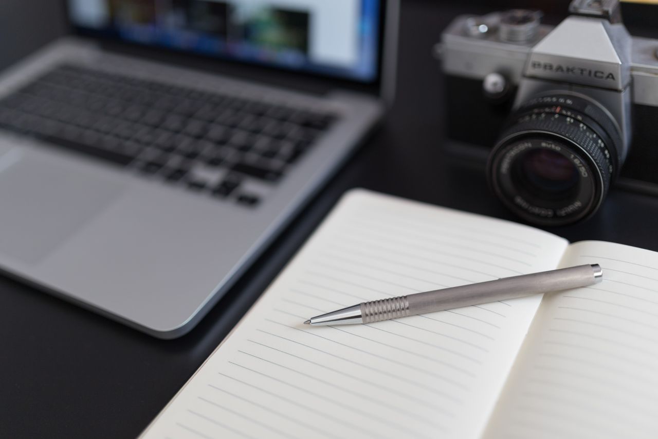 A close-up view of a laptop, vintage camera, and open notebook with a pen.