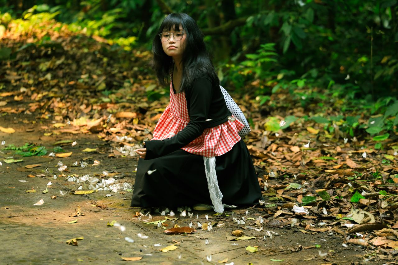 A young woman in a checkered red apron sitting on a leaf-strewn forest path in Ninh Bình, Vietnam.